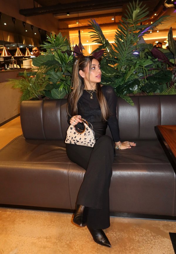 Femeie îmbrăcată în negru și cizme stă pe o bancă maro din piele, ținând o geantă cu imprimeu animalier într-un restaurant plin de plante și cu iluminare caldă.