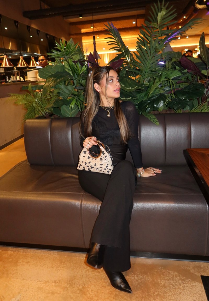 Γυναίκα με μαύρο σύνολο και μπότες κάθεται σε καφέ δερμάτινο πάγκο κρατώντας τσάντα με σχέδιο ζώου σε εστιατόριο γεμάτο φυτά και ζεστό φωτισμό.