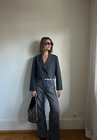 Šedý zkrácený blejzr s dlouhými rukávy, spárovaný s šedými džíny s vysokým pasem. Doplňky zahrnují černou kabelku se cvočky a tmavé sluneční brýle.