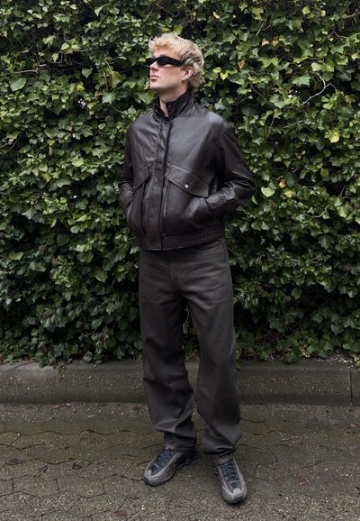 Persona rubia con gafas de sol negras, chaqueta de cuero marrón oscuro, pantalones oscuros sueltos y zapatillas grises, de pie frente a un denso muro de hiedra verde.