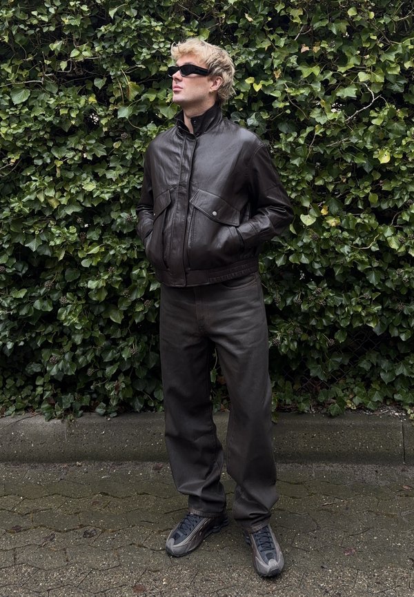 Blonde Person trägt schwarze Sonnenbrille, dunkelbraune Lederjacke, lockere dunkle Hose und graue Turnschuhe und steht vor einer dichten grünen Efeuwand.