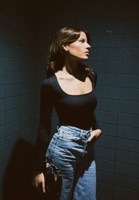 Jeune femme en haut noir à manches longues et jean taille haute, debout contre un mur sombre en carreaux, tenant un petit sac noir.