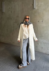 Long manteau blanc sur un pull léger, pantalon gris surdimensionné et chaussures à semelles compensées épaisses. Une écharpe grise et des lunettes de soleil complètent la tenue.