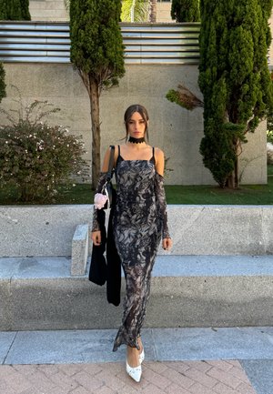 Vestido negro y gris ajustado con un patrón, manguitas largas, hombros al descubierto y una silueta de largo hasta el suelo; combinado con tacones blancos.