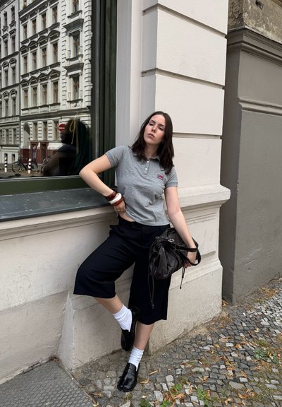 Camiseta polo gris con un logo, culottes negras, calcetines blancos y zapatos negros. Sosteniendo una bolsa negra, de pie contra una pared blanca con una ventana.