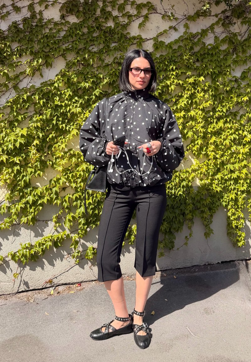 Naine kannab musta täpilise jakki, lühikesi musti pükse ja studidega musti kingi, seisab, hoides kõrvaklappe ja telefoni koos sõstrapuulehega kaetud seina taustal.