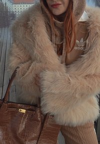 Femme portant un manteau en fausse fourrure beige et un sweat à capuche Adidas, tenant un sac à main en cuir marron texturé sur une terrasse en bois.
