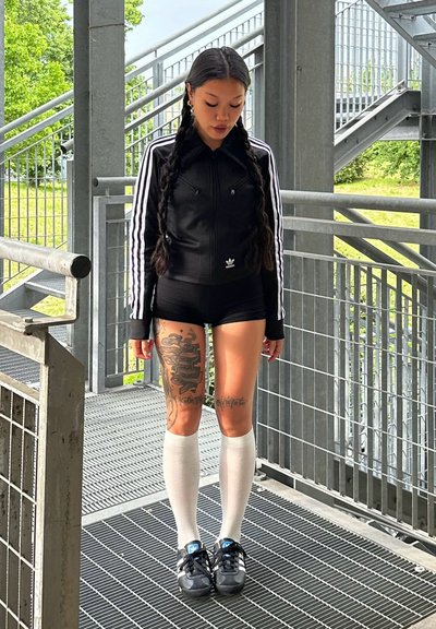 Chaqueta negra con cremallera y rayas blancas, pantalones cortos negros y calcetas blancas hasta la rodilla. Zapatillas negras con acentos azules. Tatuajes visibles en las piernas.