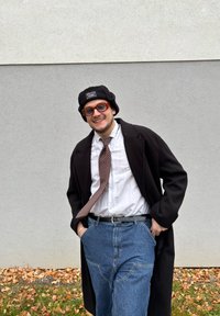 Person klädd i en svart rock över en vit skjorta och ett mönstrat slips, kombinerat med blå denimbyxor och en svart hatt, mot en grå vägg.