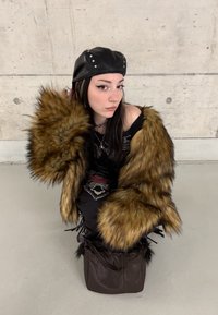 Νεαρή γυναίκα με μαύρο δερμάτινο καπέλο με τρουκς και υπερμεγέθη καφέ γούνινο τζάκετ, σκυφτή δίπλα σε τσιμεντένιο τοίχο με μια σκοτεινή τσάντα.