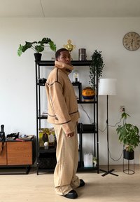 Veste en polaire beige avec col montant, détails de couture foncés, pantalon beige oversize et chaussures noires épaisses, le tout dans un intérieur moderne.
