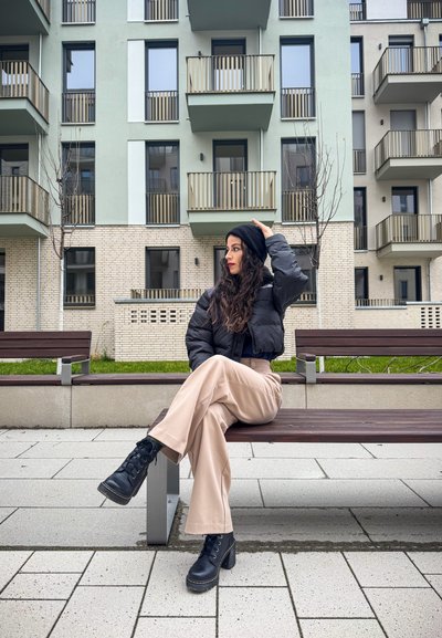 Chaqueta de plumas negra, pantalones de pierna ancha beige y botas de plataforma negras con cordones. Fondo de un edificio moderno con balcones y detalles de ladrillo.