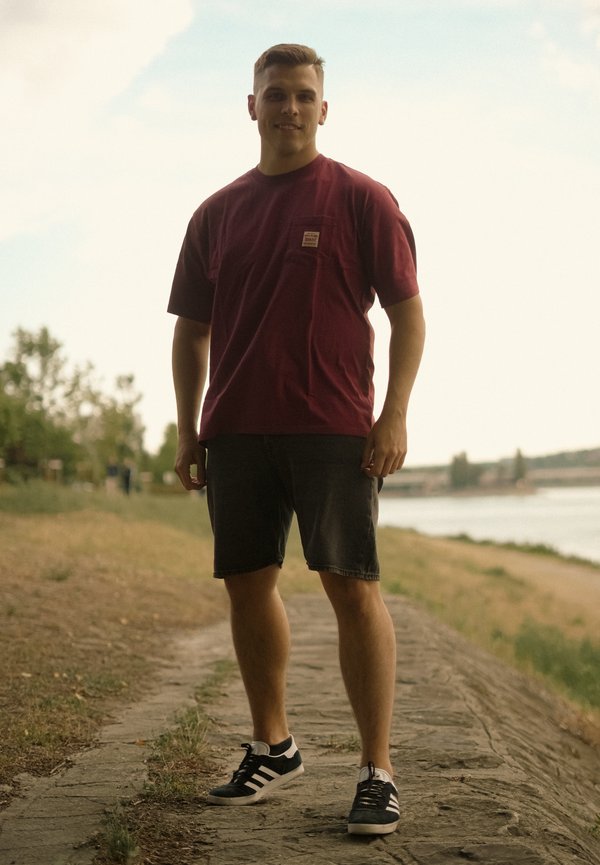 Jeune homme en t-shirt bordeaux et short noir debout sur un chemin pavé près d'une rivière avec des arbres en arrière-plan.