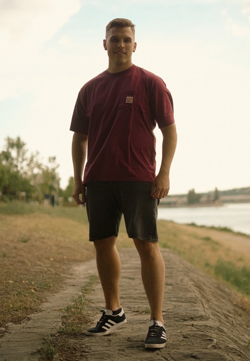 Jeune homme en t-shirt bordeaux et short noir debout sur un chemin pavé près d'une rivière avec des arbres en arrière-plan.