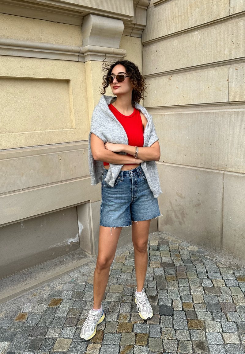 Κόκκινη στενή αμάνικη μπλούζα, ελαφρύ γκρι πουλόβερ ριγμένο στους ώμους, τζιν σορτς και γκρι αθλητικά παπούτσια με κίτρινες λεπτομέρειες, στέκεται σε πλακόστρωτο.