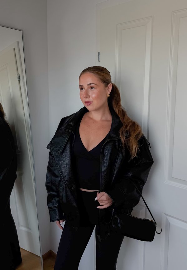Femme aux longs cheveux attachés en queue de cheval, portant une veste en cuir noire, un haut noir et un pantalon, debout à côté d'une porte blanche et d'un miroir en pied.