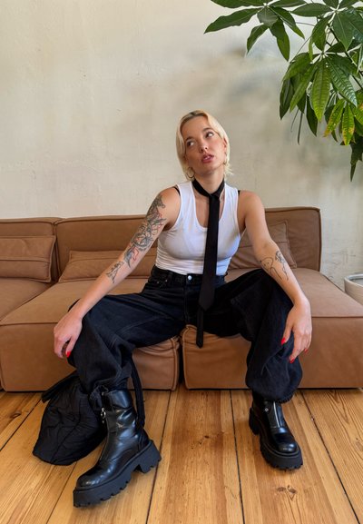 Botas de combate negras con suelas gruesas, vaqueros de denim oscuros de cintura alta, camiseta sin mangas blanca y una corbata negra. Fondo de suelo de madera clara.