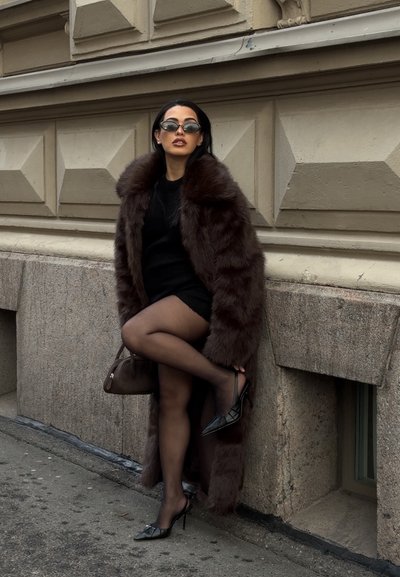 Mujer con un largo abrigo de piel marrón, vestido negro, medias transparentes, tacones negros y gafas de sol, de pie junto a una pared de piedra de un edificio.
