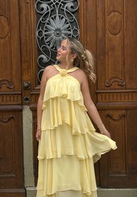 Gelbes Chiffonkleid mit geschichteten Rüschen, Halterneck-Design, Stoffblumenakzent am Ausschnitt, fließende Silhouette und leichtes Material.