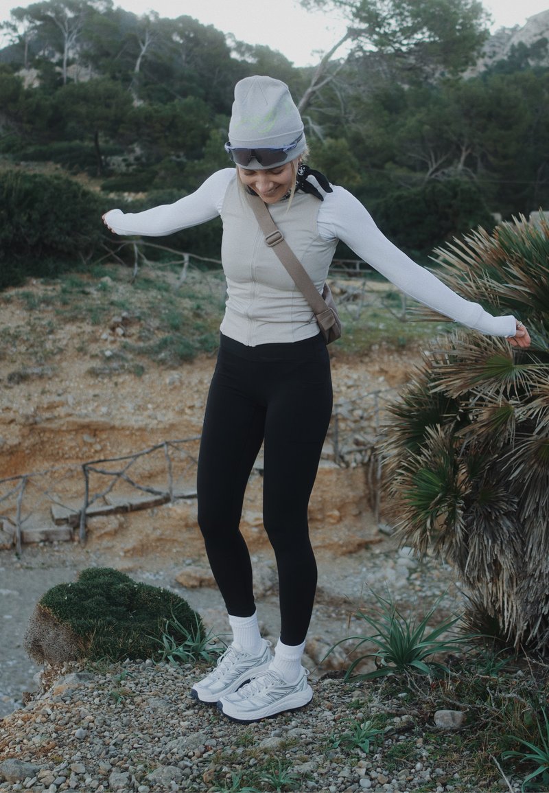 Jeune femme en tenue de sport et bonnet, en équilibre sur un terrain rocheux les bras étendus, entourée d'arbres et d'arbustes en plein air.