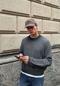 Mladic u sivoj dukserici i kapici, koristi pametni telefon, stojeći ispred teksturiranog kamenog zida na otvorenom.