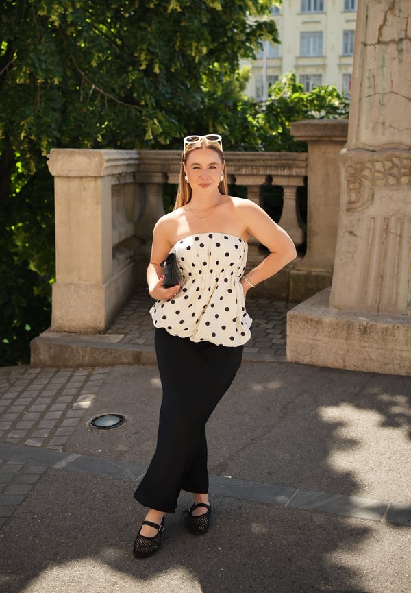 Giovane donna con top bianco senza spalline a pois e gonna nera in piedi all'aperto vicino a una ringhiera in pietra con alberi e edifici sullo sfondo.