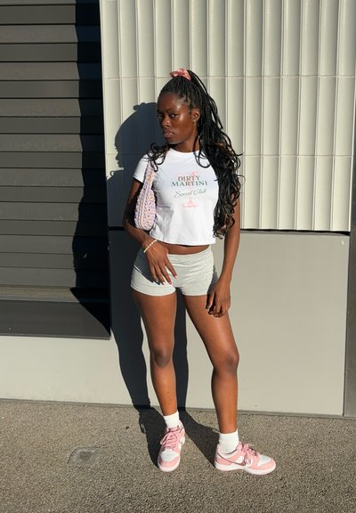 Camiseta corta blanca con el texto "DIRTY MARTINI Social Club", pantalones cortos grises, zapatillas rosas y una bolsa de mimbre rosa. Cabello largo y trenzado con un scrunchie rosa.