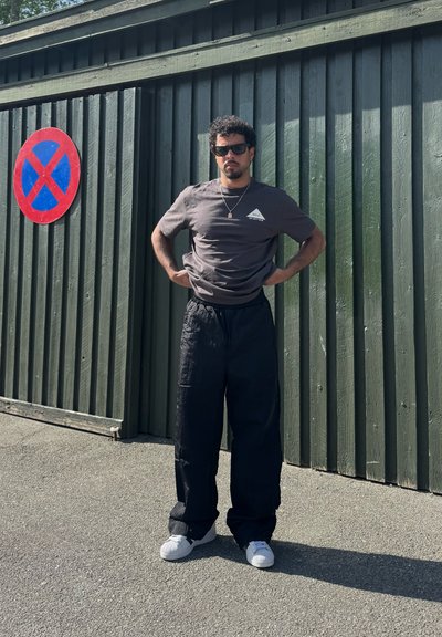 Pantalones de lino negros, camiseta gris oscuro con logotipo, zapatillas blancas y gafas de sol, de pie contra una pared de madera verde con un cartel rojo y azul.