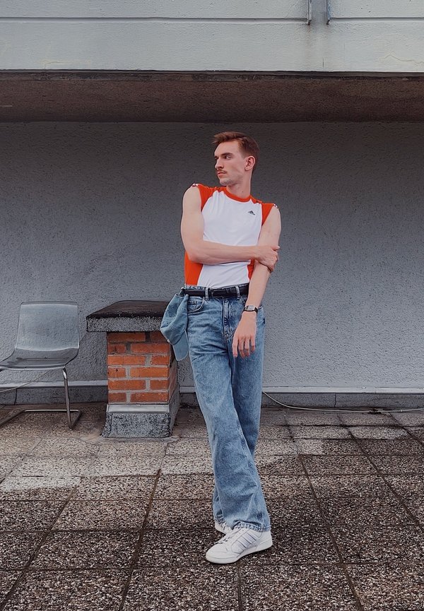 Hvit singlett med røde detaljer, lyseblå jeans, hvite sneakers. Stående posisjon på en teksturert overflate, grå vegg som bakgrunn.