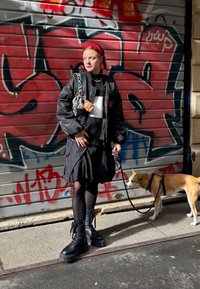 Svart bomberjakke med røde detaljer, svart skjørt, svarte strømpebukser, kraftige svarte støvler. En hund i bånd og en mønstret veske med en anheng.