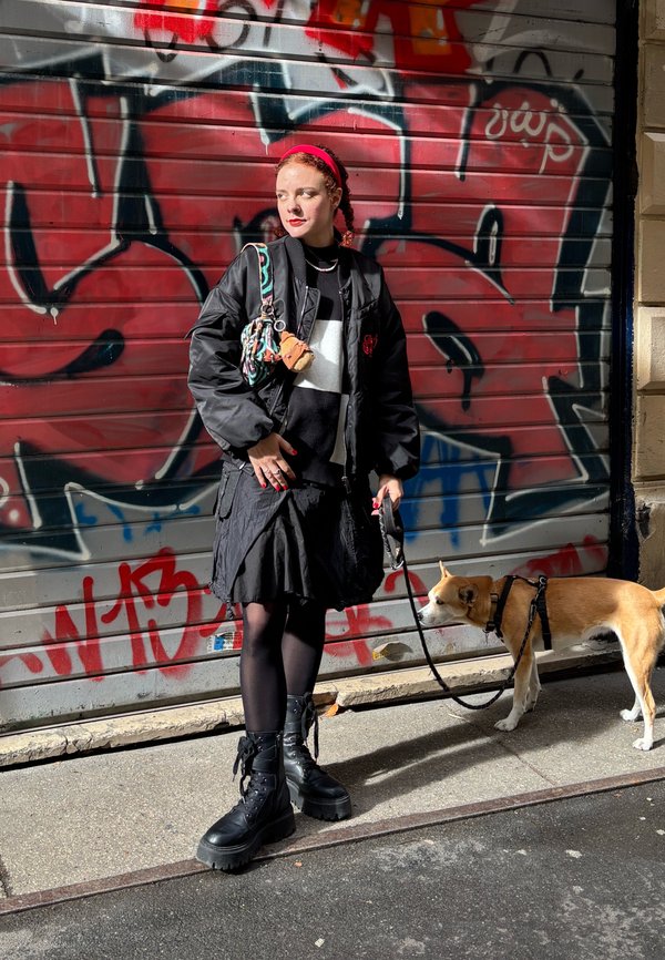 Black bomber jacket with red accents, black skirt, black tights, chunky black boots. A dog on a lead and a patterned bag with a charm.