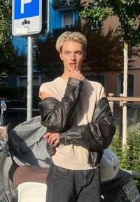 Jeune personne aux cheveux blonds portant un pull sans manches et une veste en cuir noire, debout près de scooters garés et d'un panneau de stationnement pour motos.