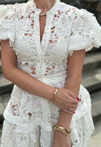 Το λευκό δαντέλας φόρεμα διαθέτει φουσκωτά μανίκια, μπροστινό κούμπωμα με κουμπιά, λουλουδάτες κοπές και στρώματα δαντέλας. Χρυσές κυματιστές βραχιόλια προσθέτουν στο στυλ.