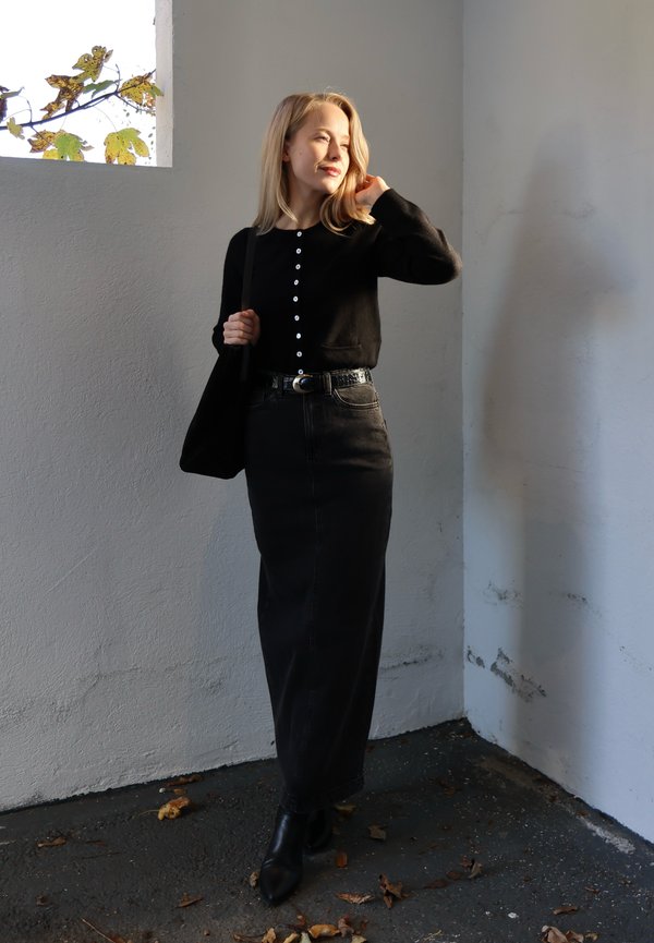 Cardigan nero con dettagli a bottoni, abbinato a una gonna lunga di denim nero. L'outfit è completato da una cintura nera e stivaletti.