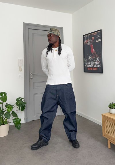 Hombre con rastas que lleva una camisa blanca de manga larga, un gorro camuflado y vaqueros holgados, está de pie mirando hacia la izquierda en una habitación con plantas y un cartel de baloncesto.