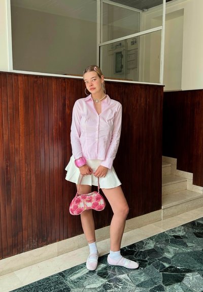 Camisa de botones rosa claro, falda mini blanca con pliegues, bolso floral rosa y calcetines rosas combinados con zapatos suaves y pálidos. Fondo de madera marrón.