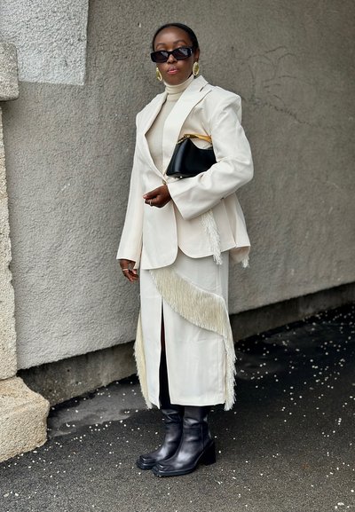 Mujer con blazer color crema y falda con flecos, botas negras, sosteniendo un bolso negro, de pie frente a una pared gris texturizada.