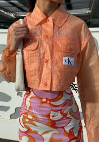 Giacca corta arancione semitrasparente con bottoni a pressione, due tasche frontali e un'etichetta con il logo Calvin Klein. Abbinata a una gonna a fantasia colorata.