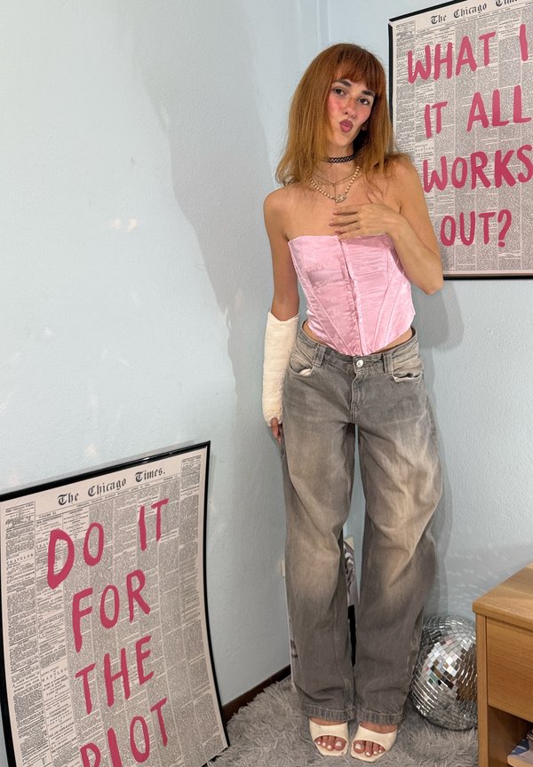 Personne avec un plâtre au bras portant un corset rose, un jean gris et des talons, se tient entre deux affiches avec un texte rose sur un mur bleu clair.