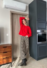 Κόκκινο πουλόβερ με πλέξη τύπου cable-knit σε συνδυασμό με παντελόνι με λεοπάρ εκτύπωση. Μαύρες μποτάκια μέχρι τον αστράγαλο. Μοντέρνο φόντο κουζίνας με ξύλινη και σκούρα ντουλάπα.