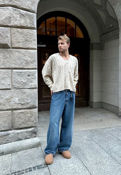 Suéter de punto color beige con escote en V y cierre de botones, combinado con pantalones vaqueros de denim azul holgados y zapatillas de piel de oveja beige.