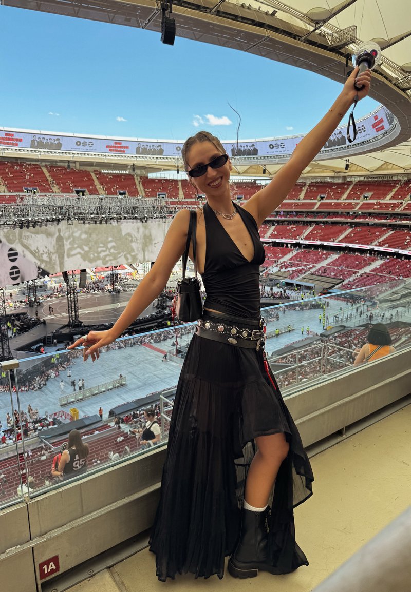 Femme en robe noire et bottes souriante et posant avec un bras levé lors d'un grand concert dans un stade en journée.