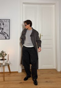 Long manteau gris charbon à épaules tombantes, associé à un pull gris clair et un pantalon cargo noir ample. Des chaussures noires à lacets complètent la tenue.