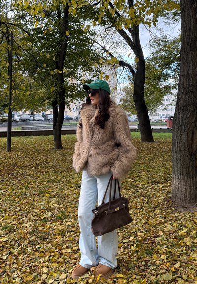 Abrigo de piel sintética marrón, jeans de color azul claro, bolso marrón, gorra verde, gafas de sol, de pie entre hojas de otoño amarillas y verdes.