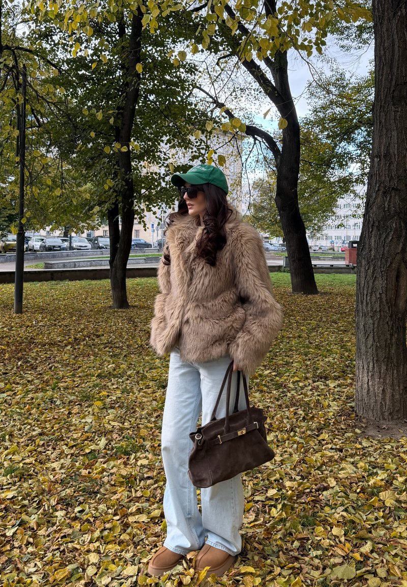 Lažna krzna rjava bunda, svetlo modri džinsi, rjava torba, zelena kapa, sunčane naočare, stoji među žutim i zelenim jesenjim lišćem.