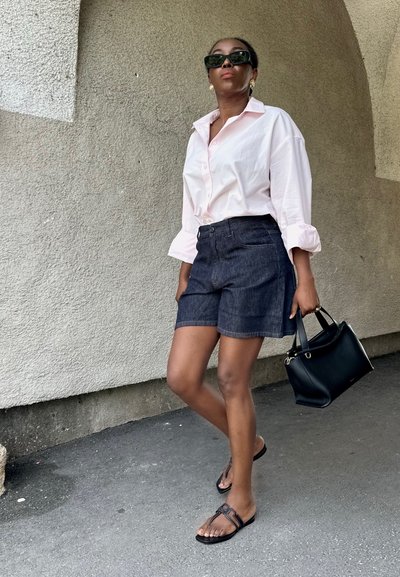 Mujer con gafas de sol, camisa rosa claro abotonada, pantalones cortos vaqueros oscuros, sandalias negras, sosteniendo bolso negro, de pie bajo un arco texturizado.