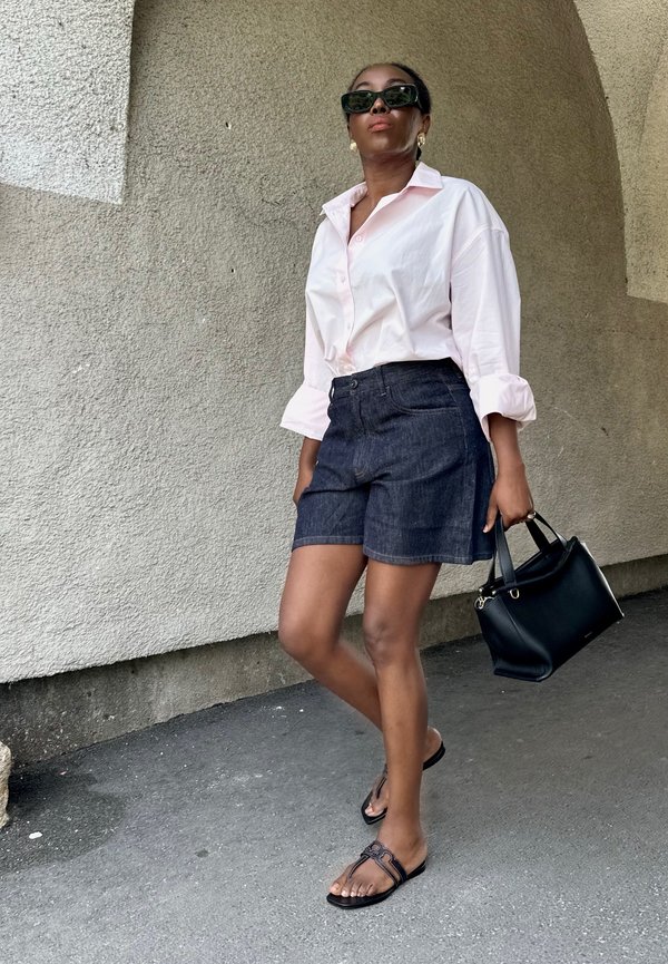 Femme portant des lunettes de soleil, chemise rose clair boutonnée, short en jean foncé, sandales noires, tenant un sac à main noir, debout sous une arche texturée.