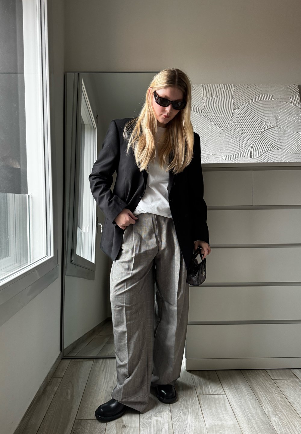 Blazer preto ajustado sobre uma t-shirt branca, combinado com calças largas de xadrez cinza e sapatos pretos robustos; o fundo espelhado reflete a luz natural.