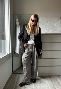Blazer preto ajustado sobre uma t-shirt branca, combinado com calças largas de xadrez cinza e sapatos pretos robustos; o fundo espelhado reflete a luz natural.