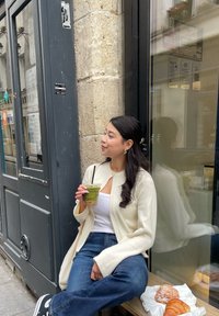 Κρέμα πουλόβερ πάνω από άσπρη μπλούζα, μπλε τζιν, κρατώντας ένα πράσινο ρόφημα. Κρουασάν και γλυκό σε χαρτί δίπλα της, γκρίζος πέτρινος τοίχος και γυάλινο φόντο.